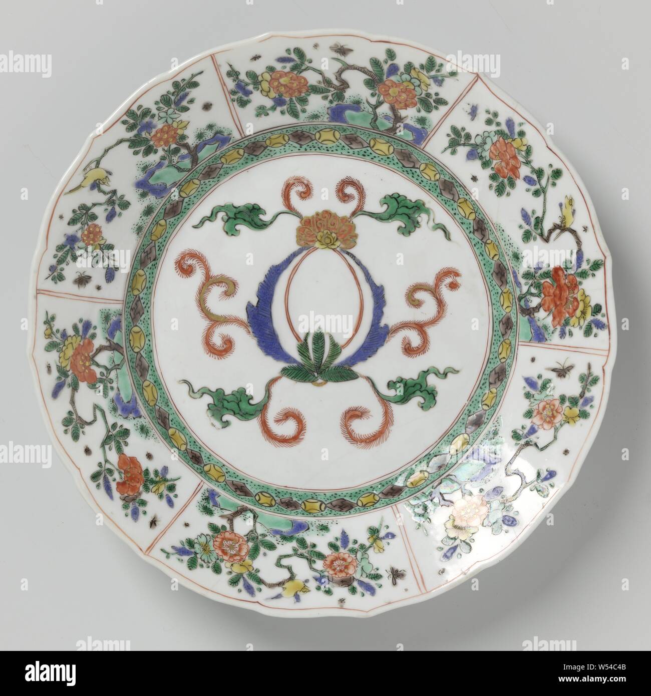 Plate with foliate scroll decoration and flowering plants, Plate with a broad, lobed porcelain border, painted in underglaze blue and on the glaze blue, red, green, yellow, eggplant, black and gold. On the shelf an empty cartouche decorated with leaf vines, the wall with a geometric motif against a green speckled ground, the border with a box decoration filled with flowering plants, birds and insects, the back with three flower sprays. Marked on the bottom with an incense burner in a double circle. Plate has been broken. Famille Verte., anonymous, China, c. 1700 - c. 1724, Qing-dynasty (1644 Stock Photo