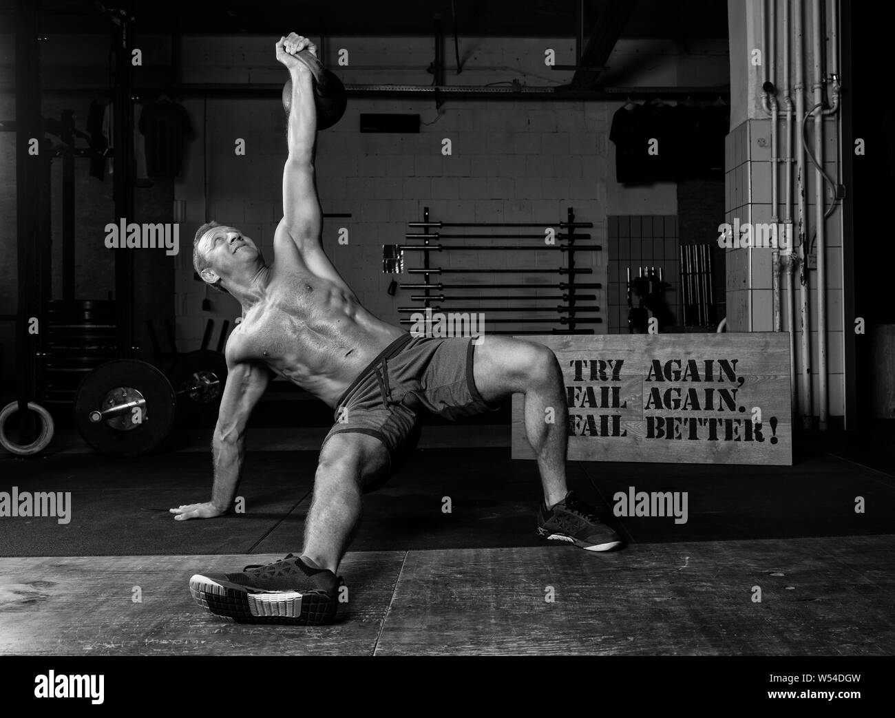 An athletic suntanned man with Sixpack is doing weight lifting with a kettlebell in a gym. Exercise Turkish get up. Black and white. Stock Photo
