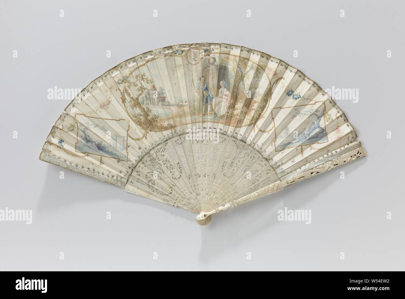 Folding fan with parchment leaf with a pastoral scene on the front and a woman under a tree on the back, on a frame of carved ivory, folding fan with parchment leaf and a landscape with a landlord and a lady in a dress cartouche. On either side of the central image a rectangle with in gouache a figure in a landscape in blue. Along the edges of the leaf are dots and stars. At the center in the back in gouache is a lady sitting by a tree. The ivory frame is partly ajour decorated, with two figures in relief, flanked by peacocks and rocaille ornaments, the back is smooth. The frame has 22 Stock Photo