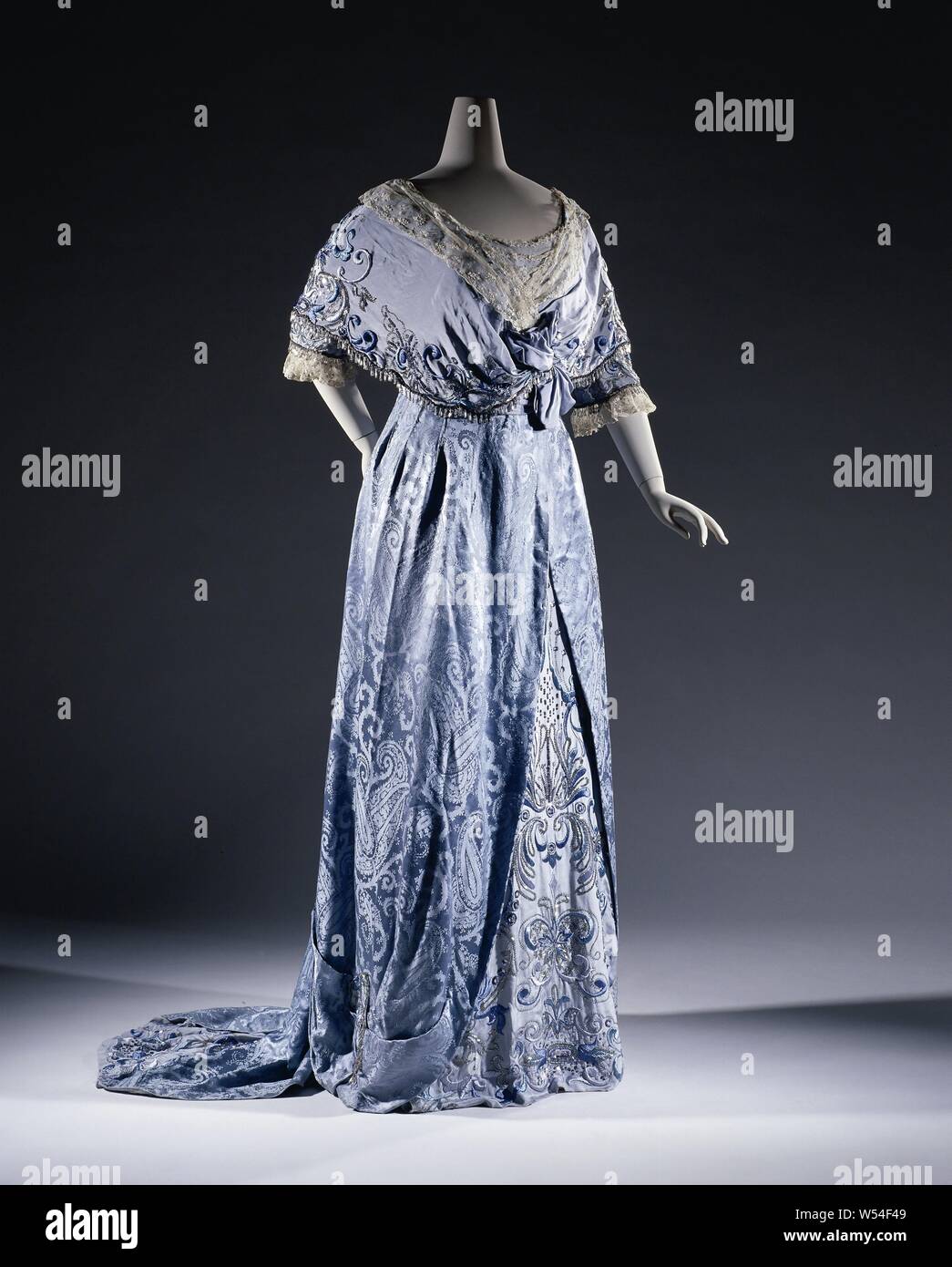 Dress in light blue silk damask with butah motifs and crêpe de chine, embroidered in two colors of blue silk, silver thread and with silver sequins, Dress with a soft blue silk damask with Kashmir palm motifs. Bodice with V-neck. Half, smooth damask sleeves and embroidered crêpe de chine. Shawl collar that runs in a pellerine. Cardigan with lace. A draped 'belt-corselet' behind. Skirt from the front and with a jacket overlay. Right below a drapery with an S-volute of the embroidered silk and bead fringe. Wrinkled from behind and open over the bottom frock, on which a triangle embroidered crêpe Stock Photo