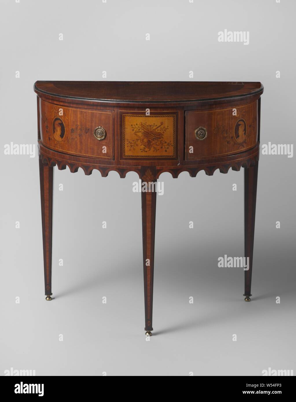 Half-table decorated with marquetry, including a trophy with musical instruments, Half-table decorated with marquetry of pink, satin and other types of wood on an oak core, on three, rejuvenating, rosewood legs and a rosette. As a rule, including a hollowed toothed rack, there are two quarter-round drawers. The line is decorated with a trophy with musical instruments and on the drawers with medallions with heads in profile, hanging on a bow and carried by flower sprays. The blade has a centrifugal veneered frame., anonymous, Netherlands, c. 1785 - c. 1800, wood (plant material), oak (wood Stock Photo