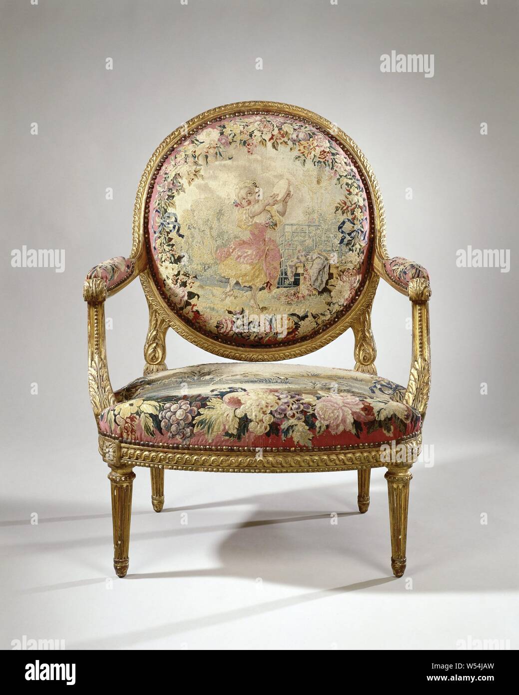 Armchair upholstered in tapestry with a dancing girl (petite danseuse) (back) and the heron's fable (seat), Armchair made of gilded beech wood, resting on conically shaped legs. The armchair belongs to an ameublement. On the pink curved seat frame and the covered oval back window, on a pink fond, representations are made within flower wreaths (tapestry de Beauvais)., Manufacture Royale des Gobelins, France, c. 1755 - c. 1765 and/or c. 1935, walnut (hardwood), gilding (material), ketting, inslag, gilding, h 101 cm × w 78 cm × d 75 cm h 43 cm × d 53 cm Stock Photo