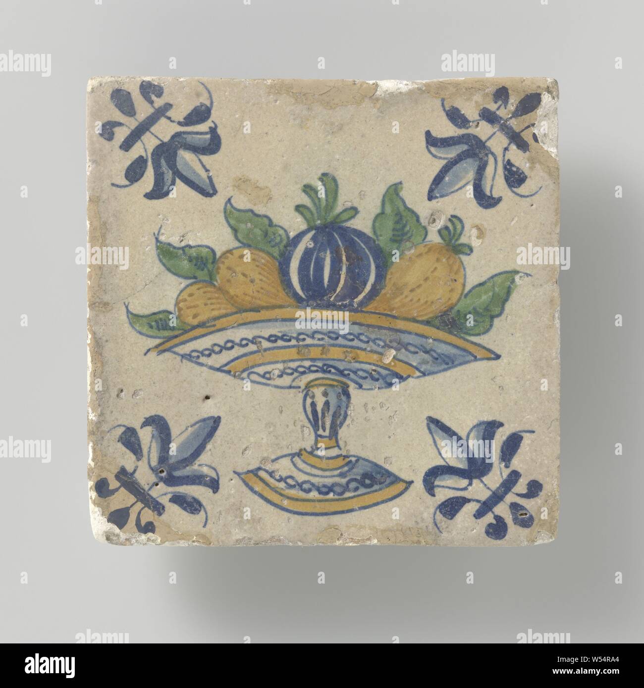 Tile with fruit bowl on foot, Tile with a multicolored (blue, green, yellow and orange) fruit bowl on foot and in the corners a lily., anonymous, Netherlands, c. 1640 - c. 1660, earthenware, tin glaze, h 13 cm × w 13 cm × t 1.5 cm Stock Photo