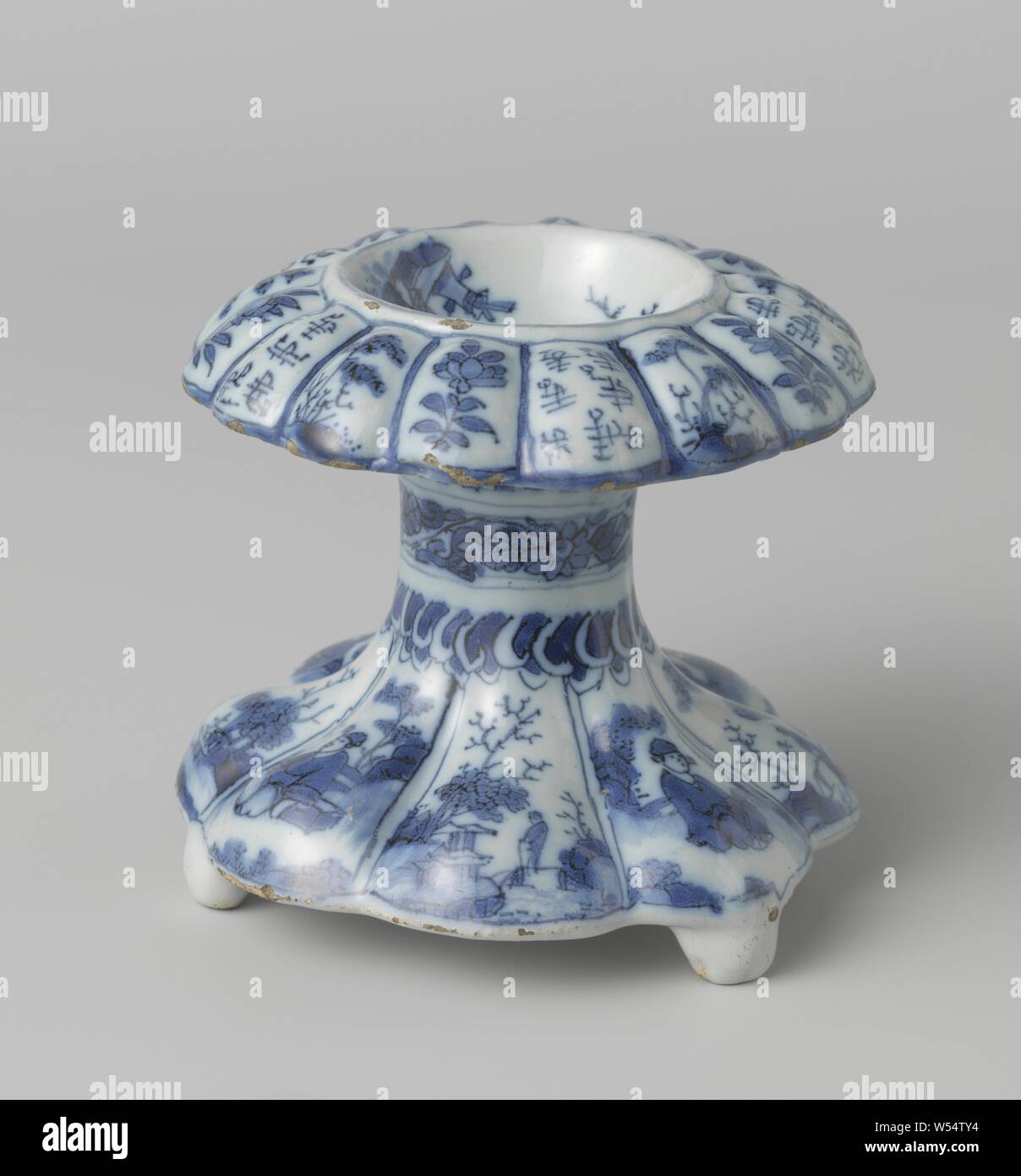 Salt cellar Salt barrel, Salt barrel of faience, pulley-shaped, on three legs, bottom part eight-lobed, the overhanging upper part is formed by a recessed round part and a sixteen-lobed edge. On the lobes of the lower part alternately large and smaller Chinese in a landscape, on the lobes of the upper part alternately twice a floral design and once Chinese characters. In the lowered part, a seated Chinese sitting by a tree. On the neck a band with flowers and leaves, above and including a floral pattern. This painting in blue with a black line on the white tin glaze. Marked., Chinese Stock Photo
