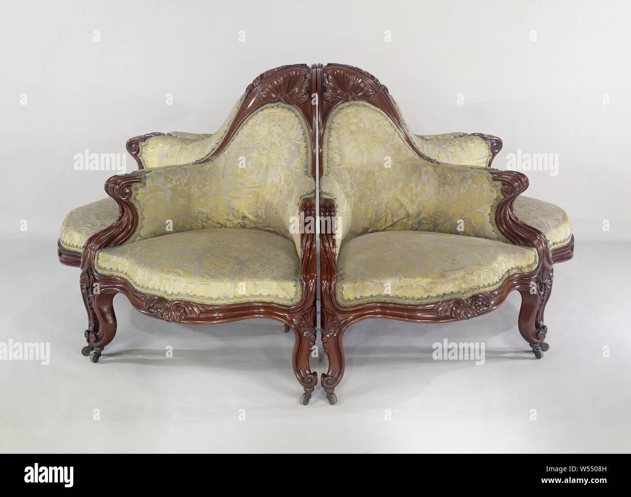 Armchair, part of an environmental divan, Armchair belonging to a set of six armchairs made of mahogany with a triangular base, forming a round sofa. Floral silk trim with trimmings. The two angled placed front legs, the rear leg, lines, struts, armrests and the crown of the backrest are bent, profiled and almost all end up in a volute. The armrests close the backrest in an arched shape and form a triangular crown that, like the fore-end and the swellings of the front legs, has shell-shaped acanthus leaves., Gebroeders Horrix, The Hague, c. 1852, wood (plant material), mahogany (wood), silk Stock Photo