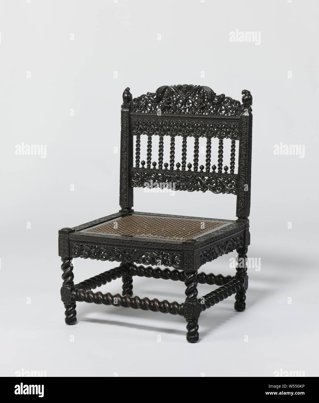 Chair with parrot-like birds on back posts, Low ebony chair with loose seat, covered with rattan wickerwork, on slung legs with houses and slung lines. The houses, front line and back styles show rosettes and tendrils. Open sculpture under the seating lines with two winged monsters and tendrils. Parrot-like birds on the back posts. Nine flung columns with leaf capitals, between which eight buds, have been placed between the sills. The chair shows sculpture with, among others, winged heads, female figures and monsters, Sri Lanka, anonymous, c. 1650 - c. 1700, wood (plant material), ebony (wood Stock Photo