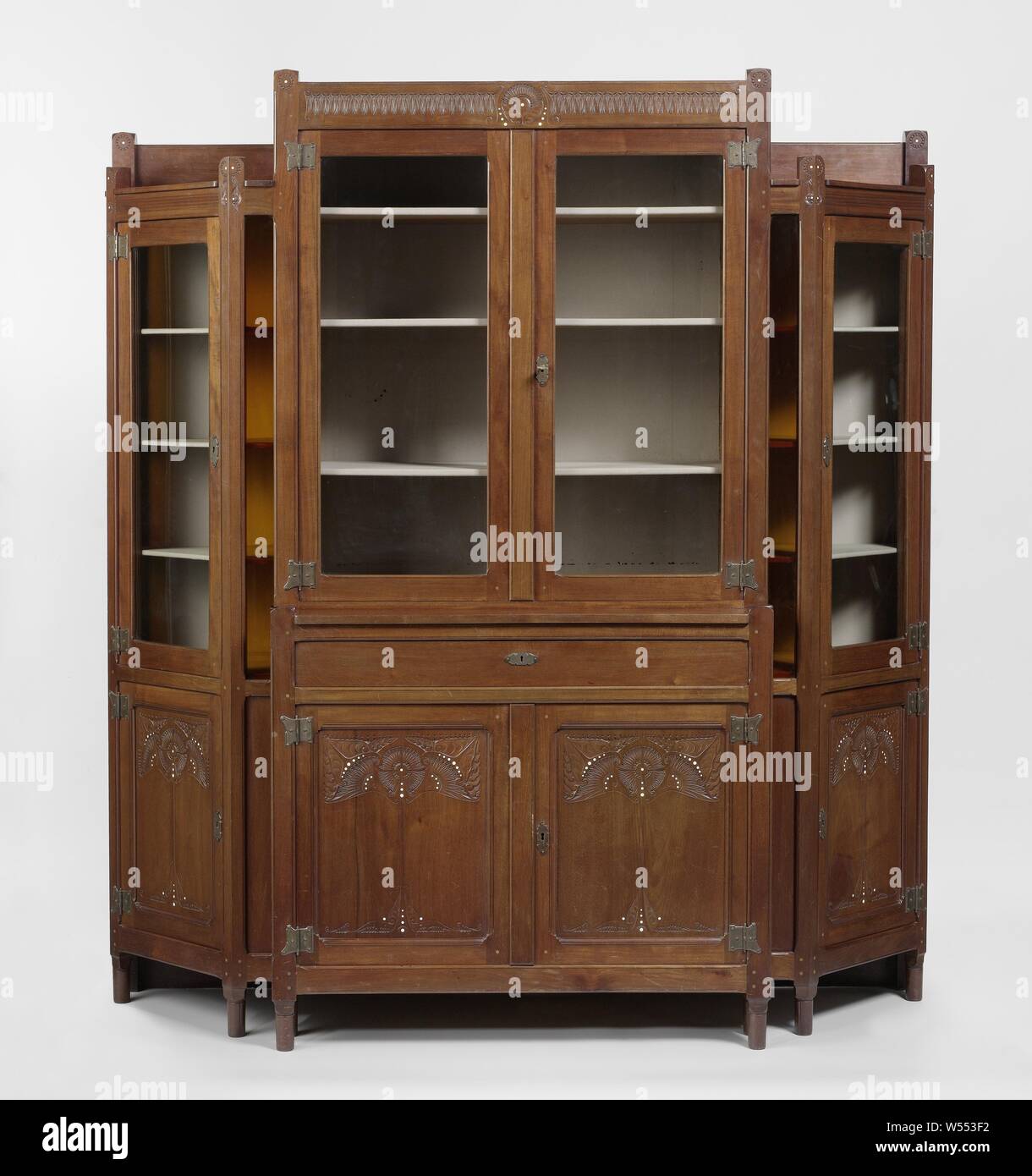 Display case consisting of a central part and two obliquely placed side parts with carved floral motifs, Display case of mahogany consisting of a central part and two angled pentagonal side parts resting on slender high legs. The middle part consists of a lower cabinet with two doors and above a drawer and a recessed upper cabinet with two high windows and a frieze. The receding side parts, which are lower than the middle part, consist of a door with a high window above them and are flanked by obliquely placed narrow parts consisting of a panel with a high window above it. There is a raised Stock Photo