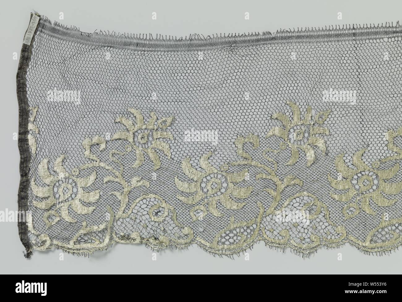 Strip of machined lace with black soil and natural-colored motifs, Strip of multicolored machined lace: machined blonde. Black soil and natural-colored motifs. The strip is probably intended or used for a frock. The repeating pattern consists of a curved branch with two openwork flowers with a wreath of crescent-shaped petals. The branch rises with a bend on the underside of the strip and bends back with a large arc where it ends with one of the flowers. The other flower originates halfway from the branch and is tilted upwards. A large cut-away leaf or reserve lies under the branch along Stock Photo