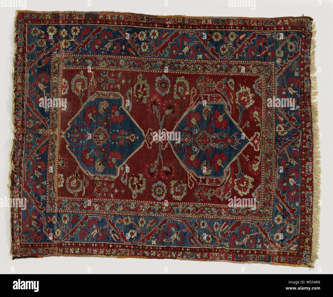 Oriental carpet, medallion rug. In the midfield on a dark terra-cotta fond are two blue leaf medallions in which kannetjew and carnations. Field filling of rosettes. Triple edge. On the wide blue main edge runs an angular edge on which a triple cluster grows., Kula, 1700 - 1900, ketting en inslag, h 155 cm × w 121 cm Stock Photo