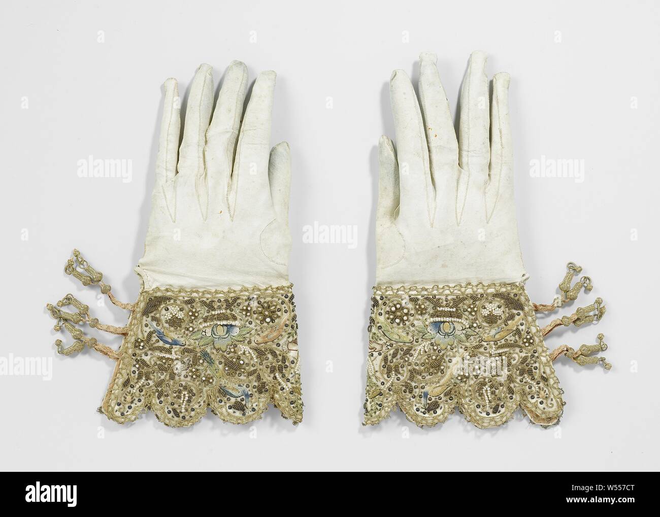 A Pair of Wedding Gloves, Left bridal glove of white wax leather, richly embroidered caps. Hooded edges. Closed with three pairs of pink lace bands, on which a gold button is slid. Symmetrical pattern of, among other things, two hands placed together in brown contour under a heart pierced with arrows and two devoted birds. Rich pattern in different colors, embroidery, pearls and sequins. Gold bobbin lace all around., anonymous, Netherlands, 1622, geheel, banden, versiering, embroidering, l 24 cm l 26 cm × w 13 cm Stock Photo