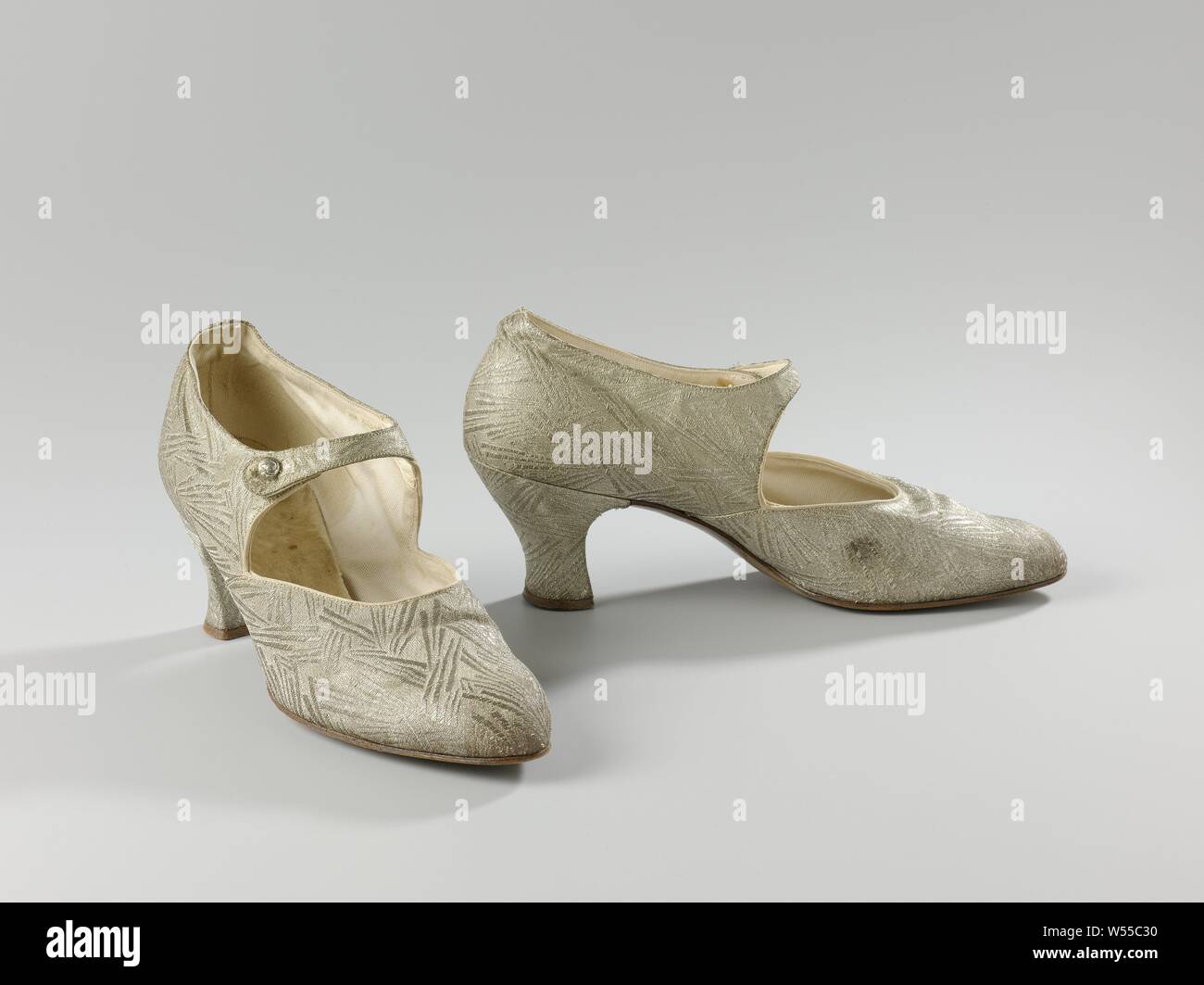 Women's shoe made of silver brocade with ankle strap, Women's shoe made of silver brocade with ankle strap. Model: The rounded pointed nose, one front sheet and two side sheets. The cut-out of the cover runs halfway down the geleng. The side sheets are perpendicular to the front sheet. An ankle strap is attached to the right-hand side sheet. A silver-colored button is attached to the left-hand side panel through a metal eye. The front of the heel runs straight down at a rounded corner. The side and back of the narrow heel are fitted and covered with silver brocade. The heel has a brown leather Stock Photo