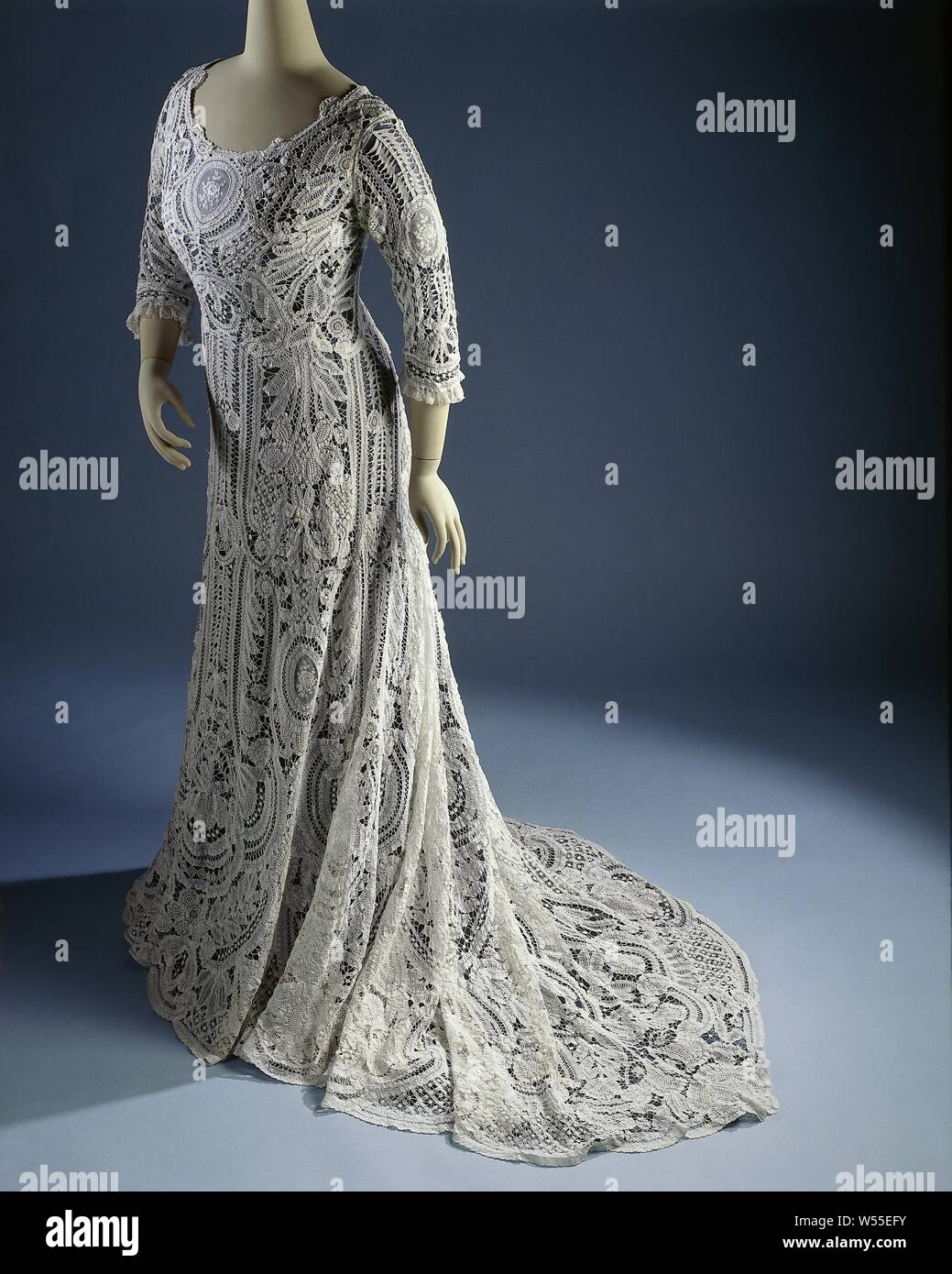 Dress with half-long sleeves and train, from cotton lace with fillings from needle lace, from white cotton lace with fillings from needle lace. The sleeves are trimmed with a strip of machined Valenciennes and with medallions from also machined lace. Design: large flowers and leaves and C-volutes, in the medallions roses. Lining of white (art?) Silk (modern)., Foliage, tendrils, branches, ornament, flowers, anonymous, Belgium (possibly), c. 1900 - c. 1910, geheel, Valenciennes lace, l 149.0 cm × l 194.0 cm Stock Photo