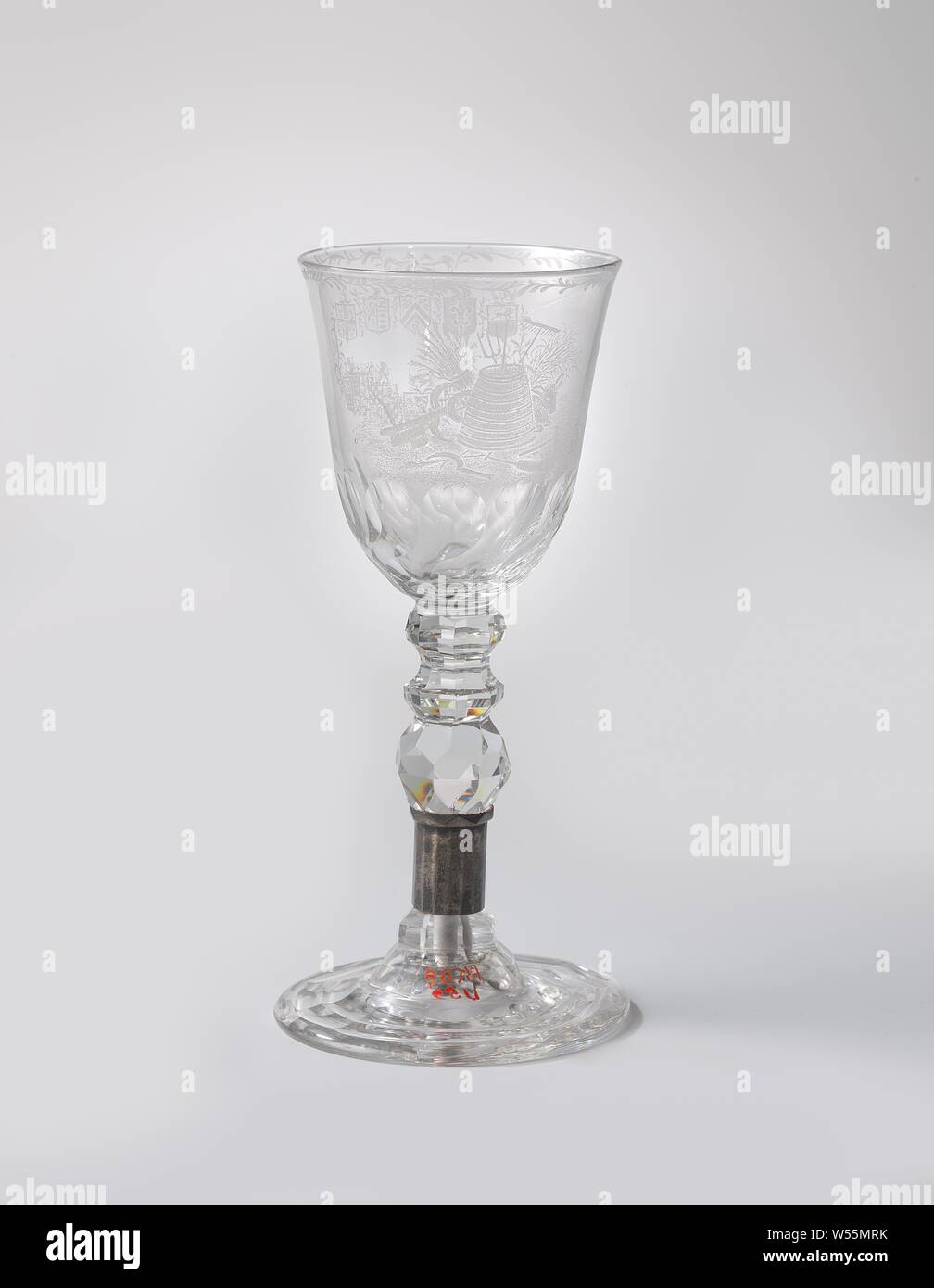 Cup with a churn in the middle of agricultural implements, Flat, facet-cut base. Baluster-shaped, facet-cut trunk with a silver frame and three knots. Conical, facet cut on the underside, rounded cup. On the chalice a pendulum of ten weapons, hanging from a bow and connected by a two branches that cross at the bottom. On the reverse, a churn in the midst of garden tools, a donkey, a grain sheaf and a bundle of hay, with a farm in the background. Along the edge a bra drink., anonymous, c. 1750 - c. 1775 and/or c. 1775 - c. 1800, glass, montuur, glassblowing, h 19.7 cm × d 8.8 cm Stock Photo