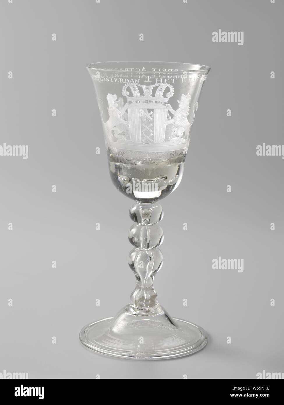 Wine glass with the arms of Amsterdam Goblet with the arms of the city of Amsterdam, curved foot with folded rim. Baluster-shaped trunk with knots and an elongated air bubble. Conical, slightly flared bowl with a thickened, rounded bottom with air bubbles. On the chalice the crowned coat of arms of the city of Amsterdam on a pedestal, on which hangs a lambrequin with frills. On the reverse, on a tiled floor, four men are seated at an oval table with an inkwell with a feather and documents. The inscription along the mouth: WELFARE OF THE WELL-TO-HAVE GOVERNMENT OF AMSTERDAM, coat of arms (as Stock Photo