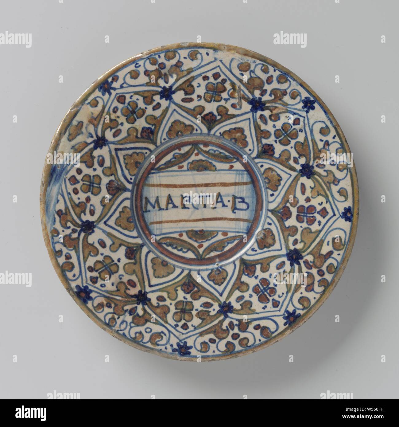 Plate, multicolored painted with round diamond-shaped compartments with leaf motifs inside and with inscription: MARTA.B, Round plate of multicolored majolica. A circle has been painted in the middle with the inscription: 'MARTA.B'. Round diamond-shaped boxes are painted around the circle, including leaf motifs., anonymous, Gabbio, c. 1530 - c. 1540, earthenware, tin glaze, lead glaze, d 22.3 cm × h 2.4 cm Stock Photo