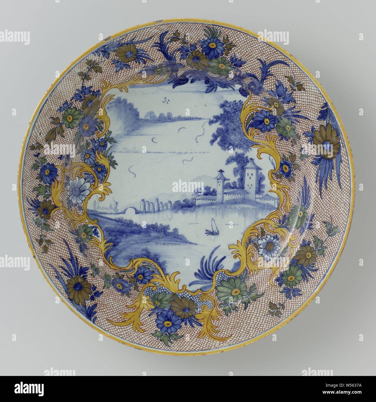 Dish painted with a river landscape, Dish of faience. On the shelf a landscape in blue. Around it a multicolored flower border., anonymous, Delft, c. 1750 - c. 1775, d 35.2 cm × h 4.8 cm Stock Photo