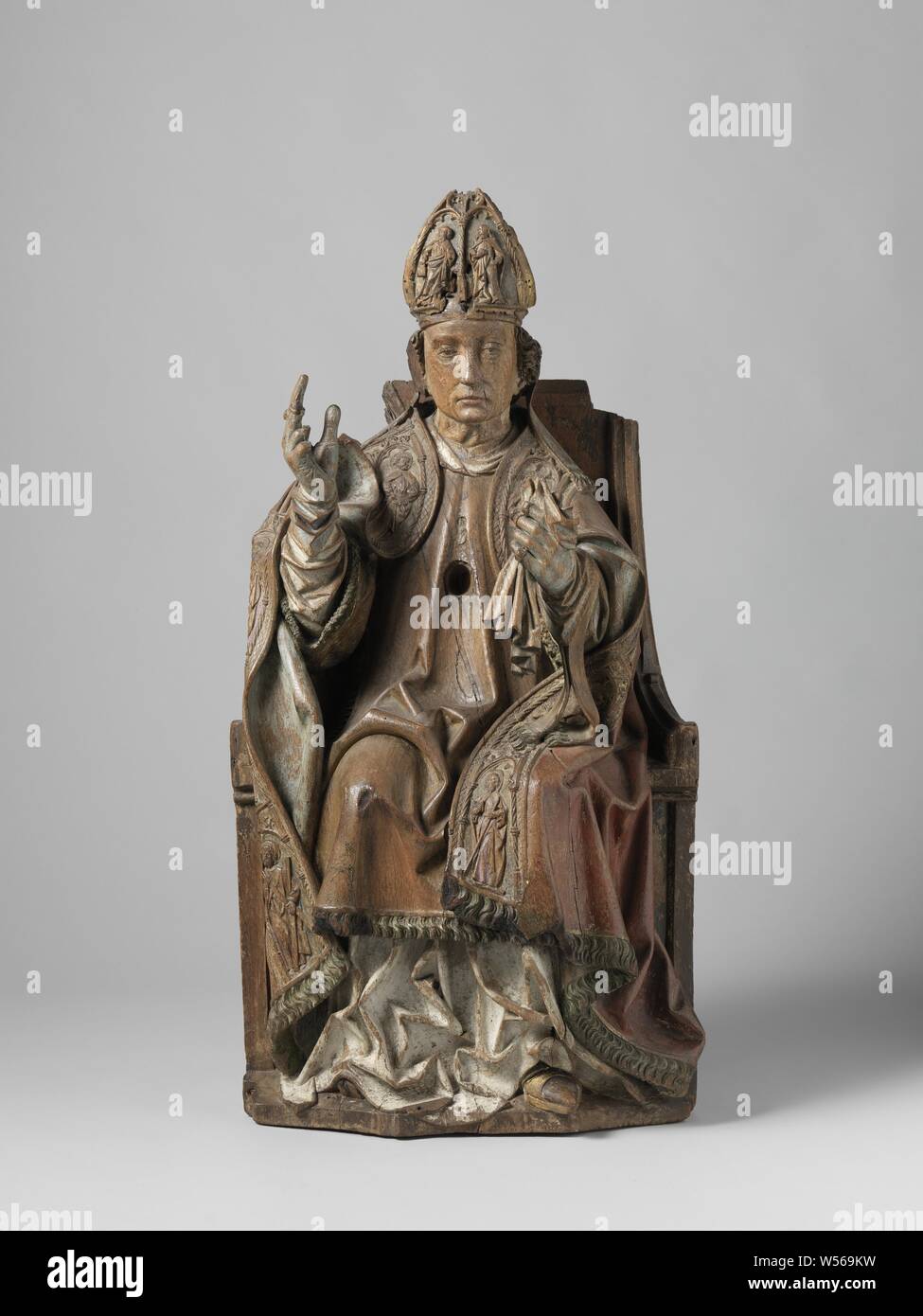 Enthroned bishop, Holy bishop sits on full throne and looks straight ahead. Right hand makes a blessing gesture, in the left hand he held the curve with the sudarium. He wears a bar, amict, albe, dalmatia and choir hood with aurifrisia with apostle figures. The choir hood curls up above the right arm. Miter with two figures and ribbons up to the shoulders. Over the left wrist he wears a manipel and further gloves over which rings. In chest oval cavity with glass., anonymous, Nederrijn, c. 1490, oak (wood), h 96.0 cm × w 50.2 cm × d 40.3 cm Stock Photo