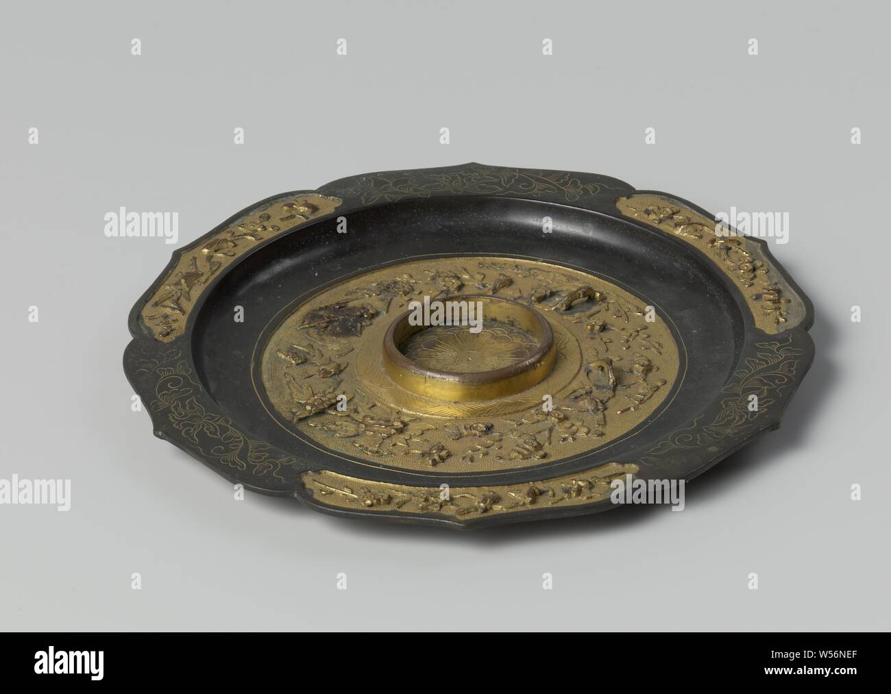 Saucer, Dish of fire-gilt suassa, with a scalloped brace-shaped rim and in the middle an upright ring, inside which a cup fits. The border is black-enamelled, with three carved floral motifs and three cartouches (medallions) with embossed gold-plated decorations of branches with flowers and birds. On the bottom, around raised ring (with weld seam), black-enamelled band with carved stripe pattern, inside gilt relief cut out of branches with leaves, flowers and birds. Carved inside raised ring image of an opened flower. The underside of the dish is smooth, Vietnam, Canton, Dutch East India Stock Photo