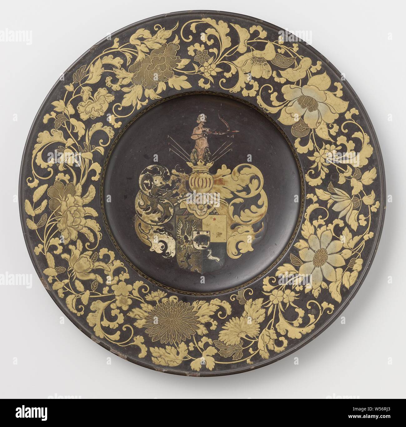 Decorative Dish Lacquerware wall plate with coat of arms Huydecoper, Plate with gilded lacquerware and colored lacquerware on black ground. The flat border is decorated with flowers and leaf vines in gold lacquer. In the middle the Huydecoper weapon with helmet and archer as helmet sign. The figure, turned to the right, is pink, bow and arrow show remnants of red. Around the shield and helmet arcanthus leaves in gold, dark gray and dirty white. The reverse side of the plate is orange, Joan Huydecoper van Maarseveen (I), anonymous, Japan, c. 1650 - c. 1660, wood (plant material), lacquer Stock Photo