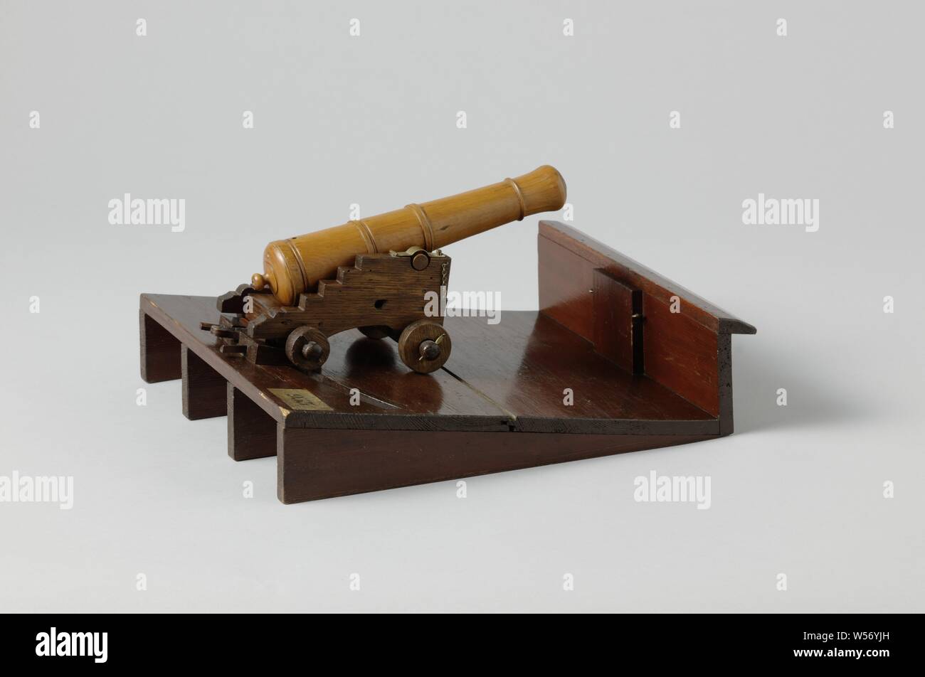 Model of a Gun and Carriage on a Ship Deck, Model of a gun on a horse on a sloping deck with board on the low side. The wooden barrel is 26 cm long. The rolling horse, narrower at the rear, consists of two cheeks with four steps and an arched cut-out at the bottom, which are connected by the calf, a breast piece, a crossbar in the middle, the soleplate and a bar under the soleplate, and the ashes. The adjusting wedge is missing. The wheels are made of wood and the front wheels are larger than the rear wheels. There is a mechanism in the deck above which hooks under the rear axle body of Stock Photo