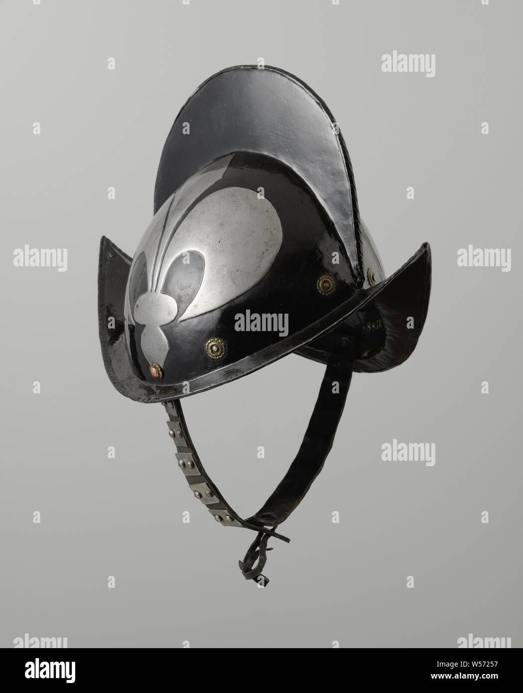 Morion helmet with extruded lily, Morion helmet, iron, blackened with white extruded lily, made from two halves. High helmet ball with high comb whose edge is cabled. Eight yellow copper rosettes surround the base of the helmet ball. The flap has high rising points on the front and back. The edge of the valve is flanged. On the left and right side on the helmet ball copper-colored rivets with which the chin strap is attached on the inside. The leather chin strap consists of two leather straps whose ends are attached to each other. Each strap has five metal plates to which two rivets are Stock Photo