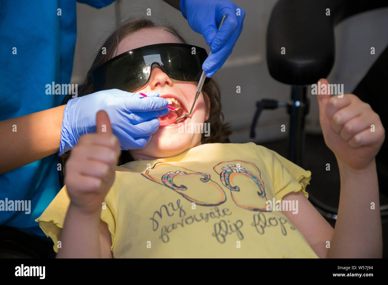 Young girl 7 yr / seven year old child receiving a fluoride coating (preventative treatment) on her teeth, wearing protection sun glasses and giving thumbs up during check up at an NHS dental practice / surgery. UK. (111) Stock Photo