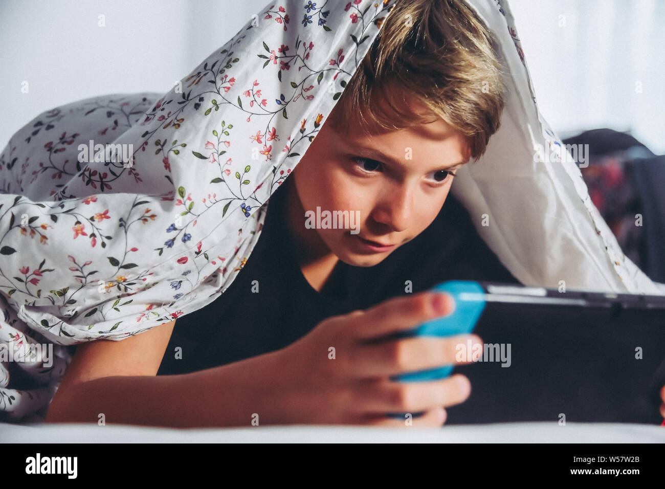 Young boy playing video games with his console, impersonating himself in the game having fun with a new game. Child using console leaning in bed in a Stock Photo