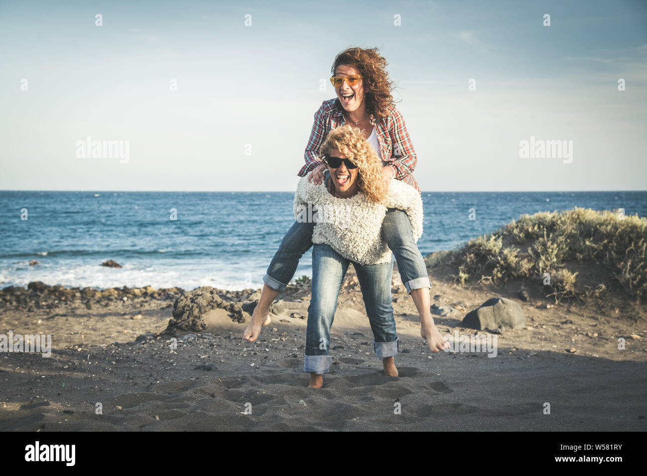 Portrait of two pretty woman enjoy free time. Smiling middle age girls giving her laughing friend piggyback while enjoying the day together at the bea Stock Photo