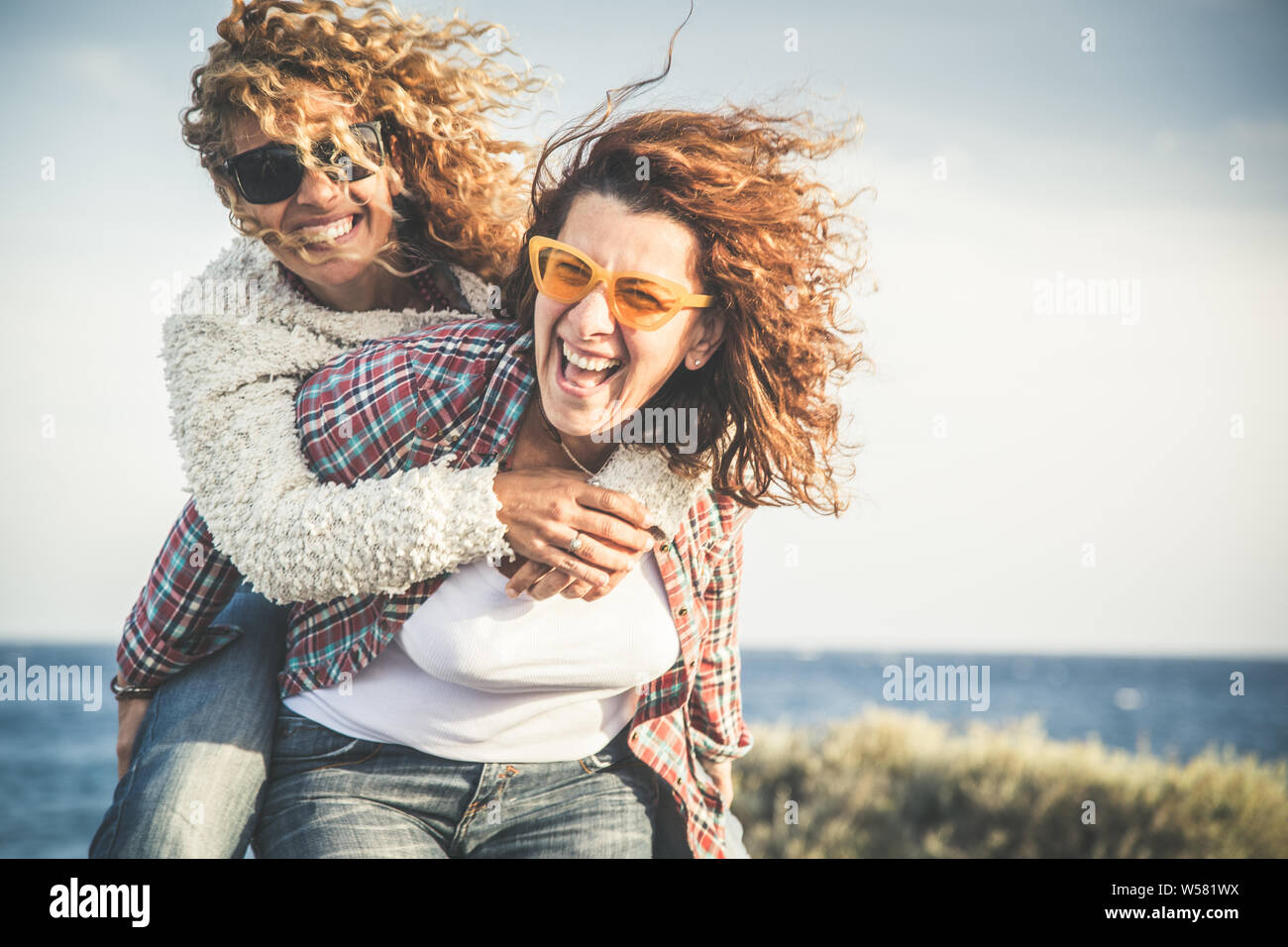 Portrait of two pretty woman enjoy free time. Smiling middle age girls giving her laughing friend piggyback while enjoying the day together at the bea Stock Photo