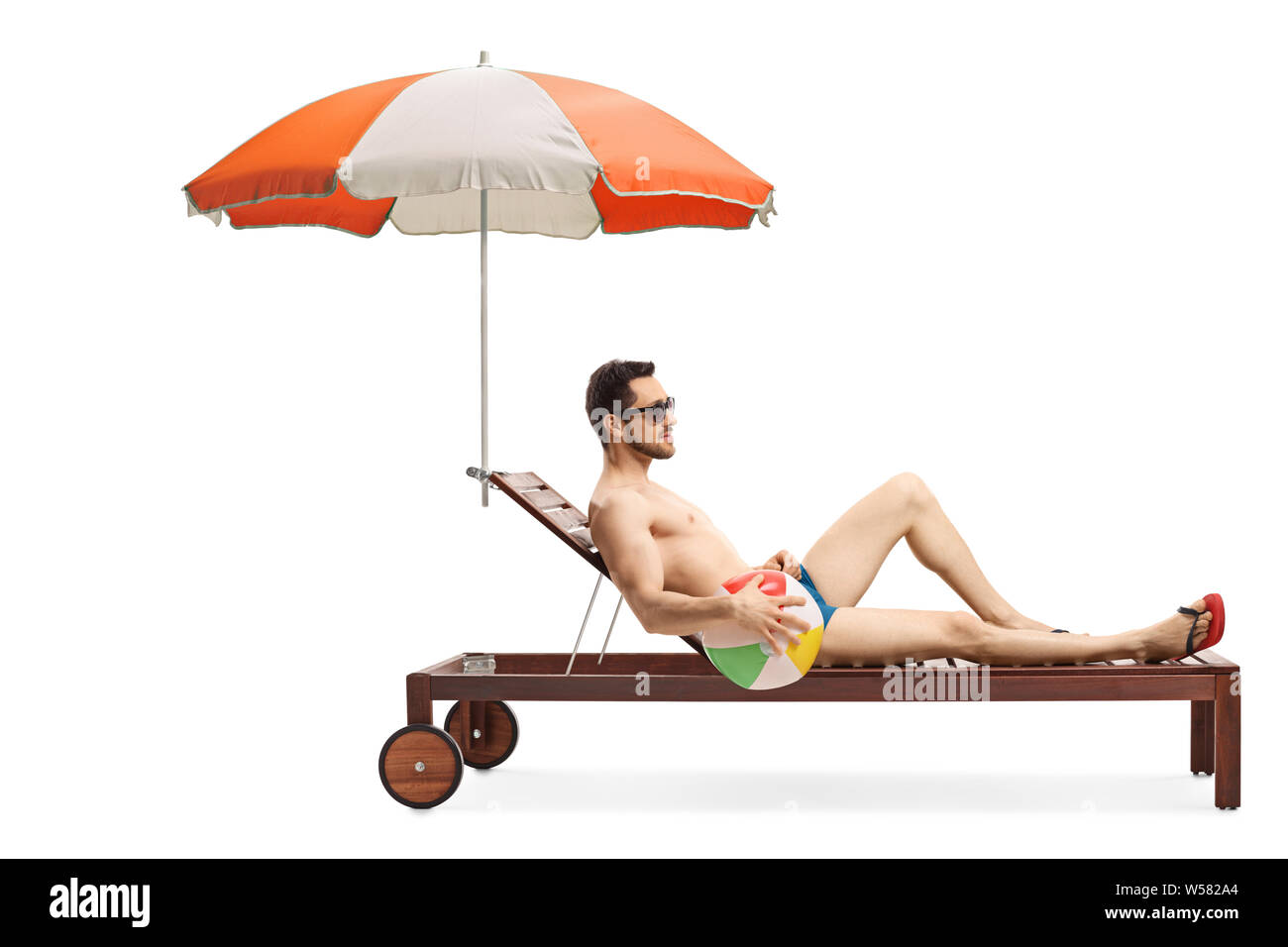 Full length shot of a young man on a sunbed with an umbrella holding an inflatable ball isolated on white background Stock Photo
