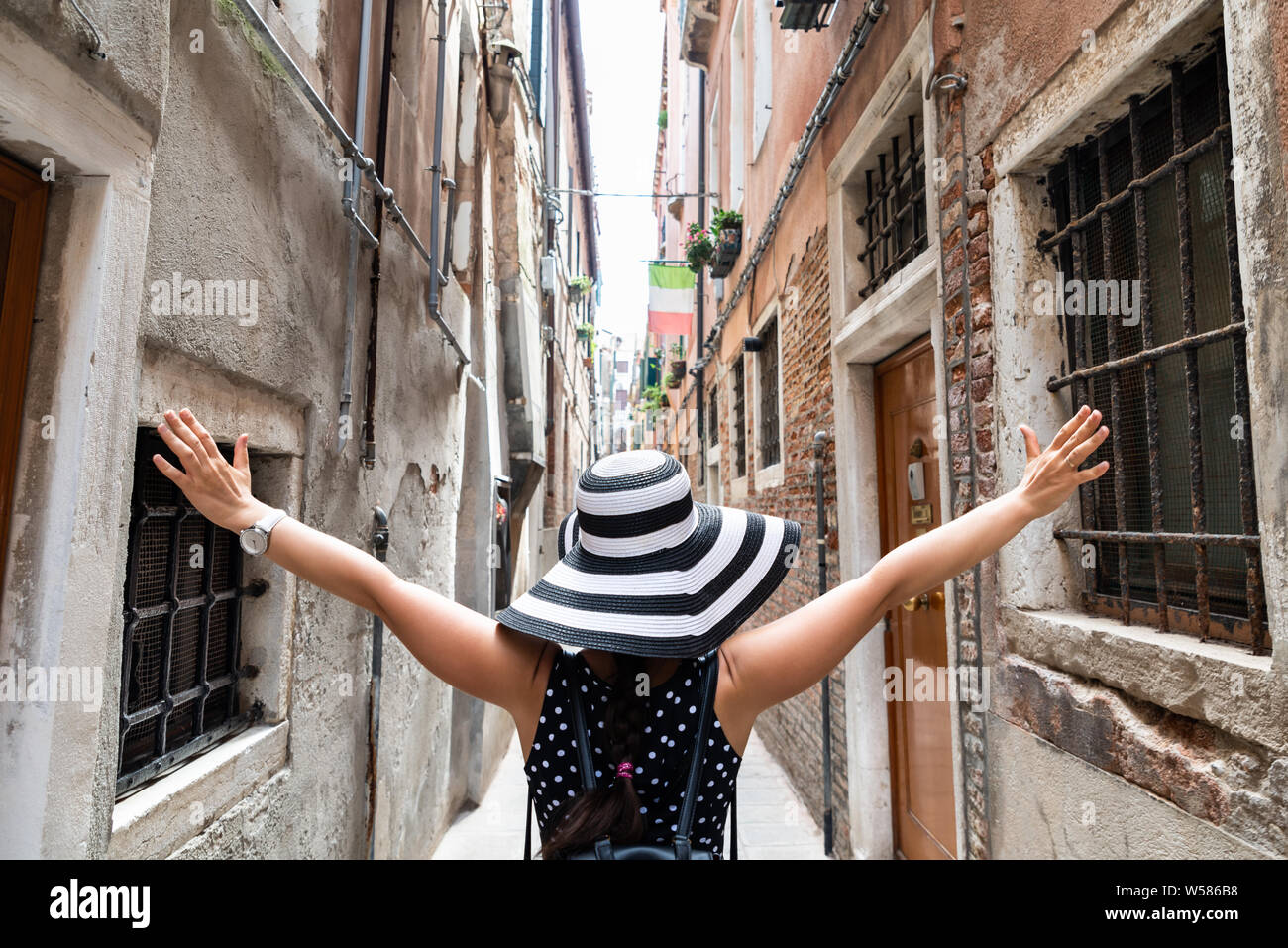 Rear View Of Woman Wearing Stripes Hat With Her Arms Outstretched Standing In Narrow Alley Stock Photo