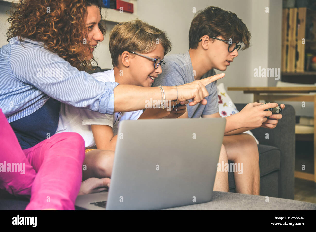 Family sitting on sofà at home using game console. Mother smiling with two young brothers playing video game. Gaming on line with friends anywhere. Be Stock Photo