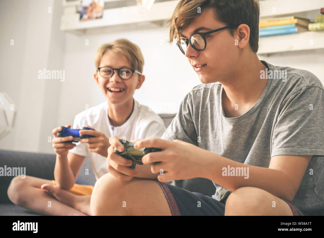 Young brothers enjoy video game at home. Smiling kids playing with console sitting on a sofa Teenager and brother playing with wireless remote control Stock Photo