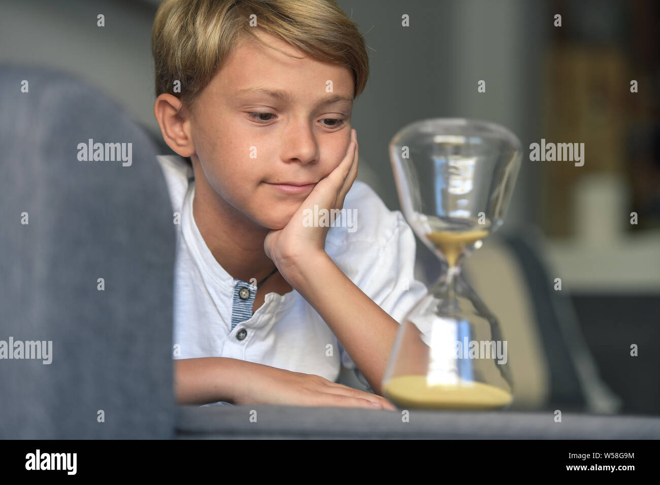 Handsome blond smiling guy lying with his hands under his face looks dreamily fascinated slow-moving sand in glass hourglass. rapt gaze, concept of de Stock Photo