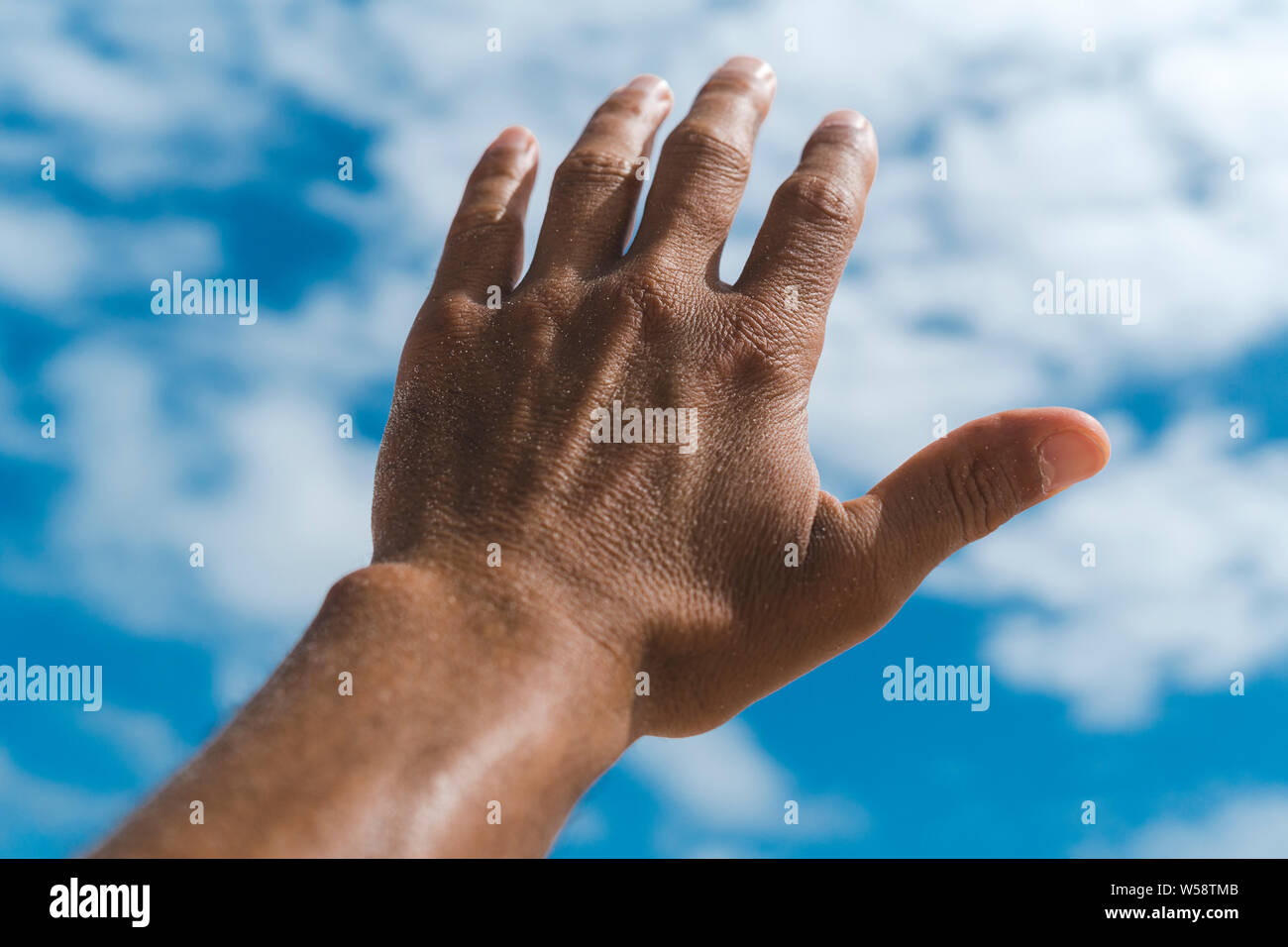 Sun tanned hand of hispanc male against blue sky with clouds Stock Photo