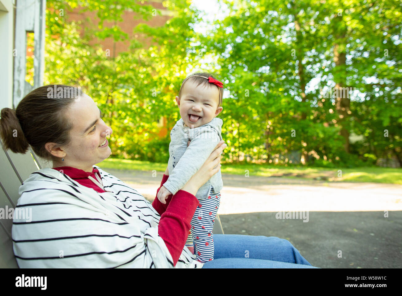 Baby girl standing on mother's lap looks at camera sticking tongue out Stock Photo