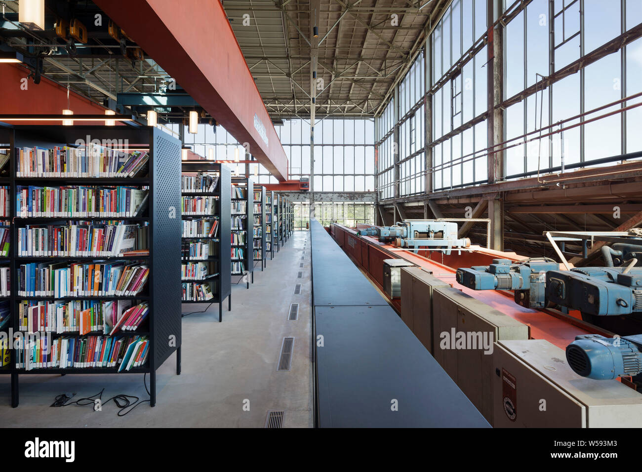 Bookshelves on upper level, with orange industrial gantry overhead and orange and blue industrial machinery to right. Bibliotheek LocHal, Tilburg, Net Stock Photo
