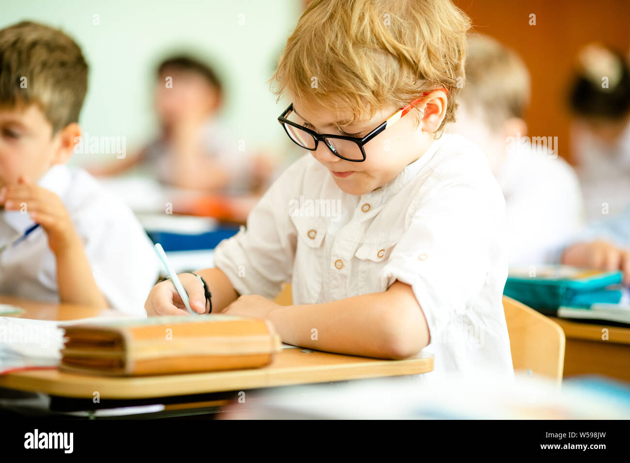 Happy Cute Clever Boy Is Sitting At A Desk In A Glasses With