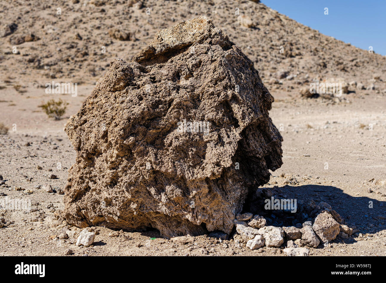 Closeup of large desert boulder showing textures and details of rock. Boulder is setting on rocky ground with barren desert mountain beyond under blue skies. Stock Photo