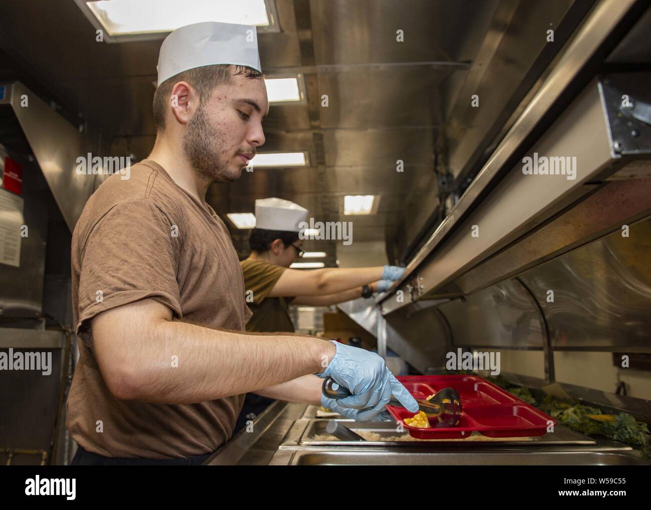 190725-N-KG461-1053  ARABIAN GULF (July 24, 2019) Operations Specialist 3rd Class Mario Cortez, from La Puente, Calif. assigned to amphibious assault ship USS Boxer (LHD 4), serves food to Sailors and Marines in the mess deck galley, July 25, 2019. Boxer is part of the Boxer Amphibious Ready Group and 11th Marine Expeditionary Unit and is deployed to the U.S. 5th Fleet area of operations in support of naval operations to ensure maritime stability and security in the Central Region, connecting the Mediterranean and the Pacific through the Western Indian Ocean and three strategic choke points. ( Stock Photo