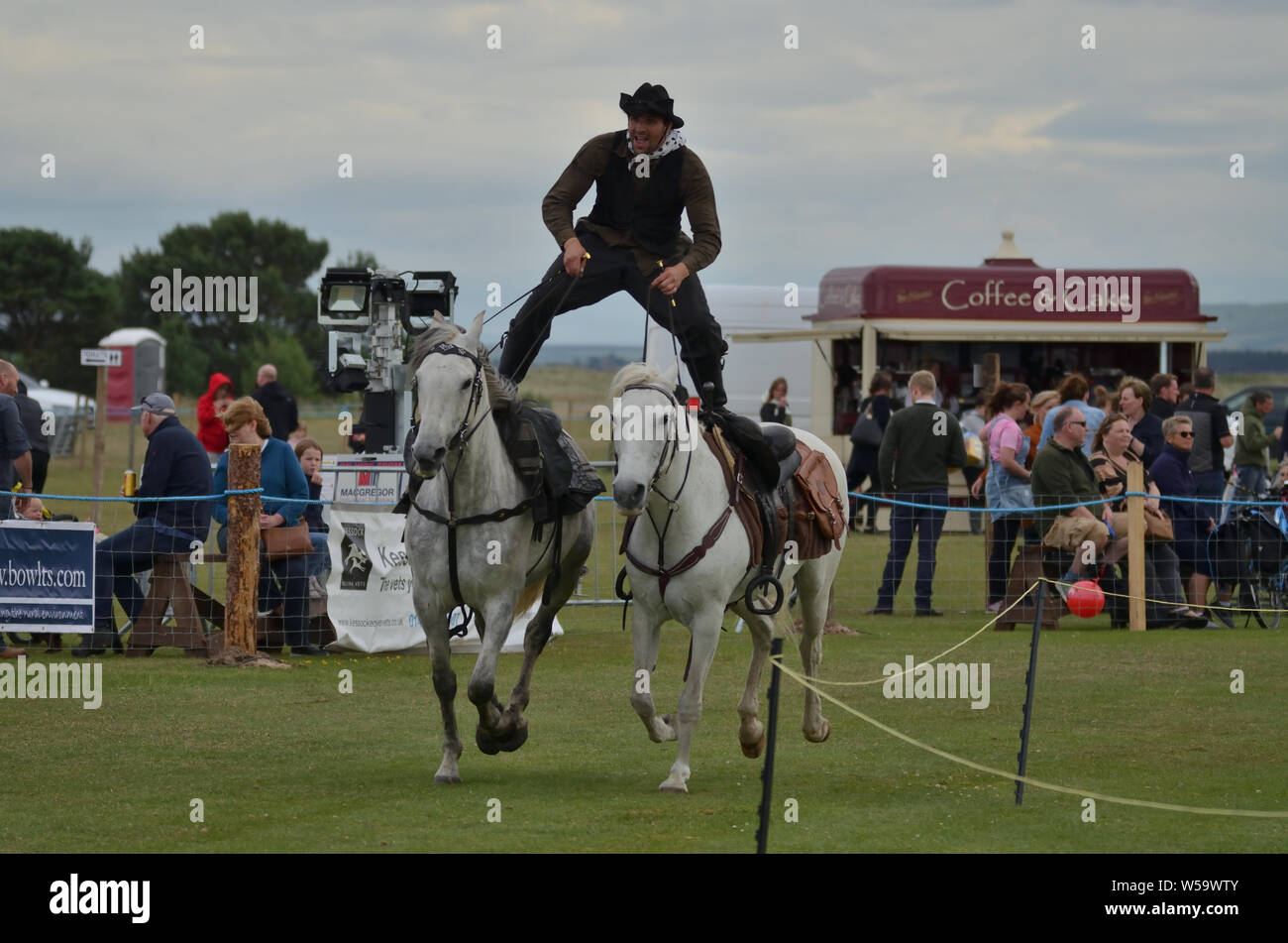 A man in Cowboy dress riding astride on two horses as part of a Wild West horse display at the 2019 Sutherland County Agricultural Show, Scotland, UK Stock Photo