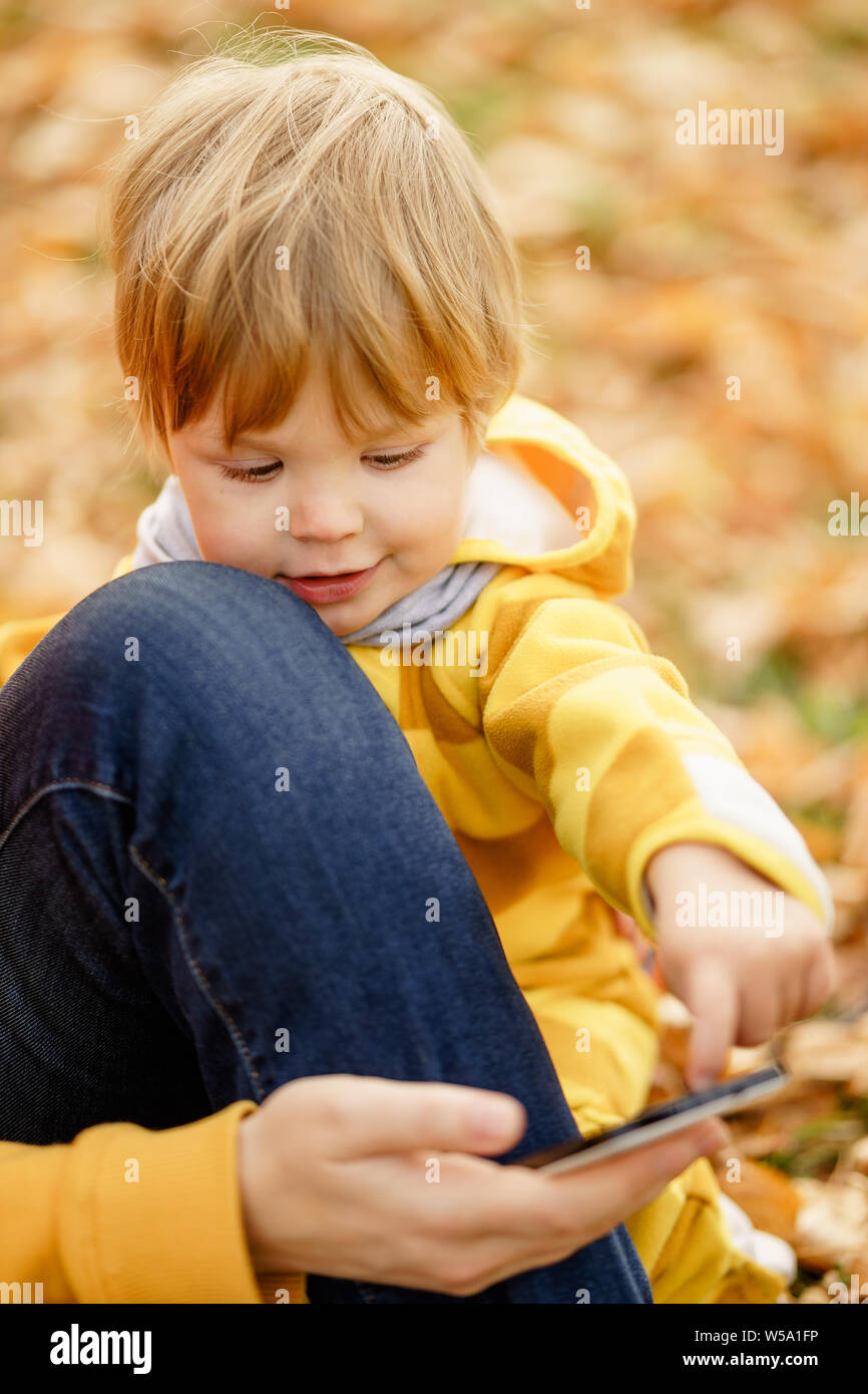 Concept: family, kids. Happy little child, baby boy laughing and playing with mother's smartphone in the autumn on the nature walk outdoors at park Stock Photo