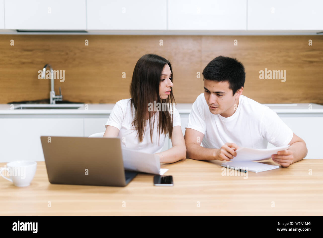 Young couple is sitting at kitchen table, use laptop and taking care of