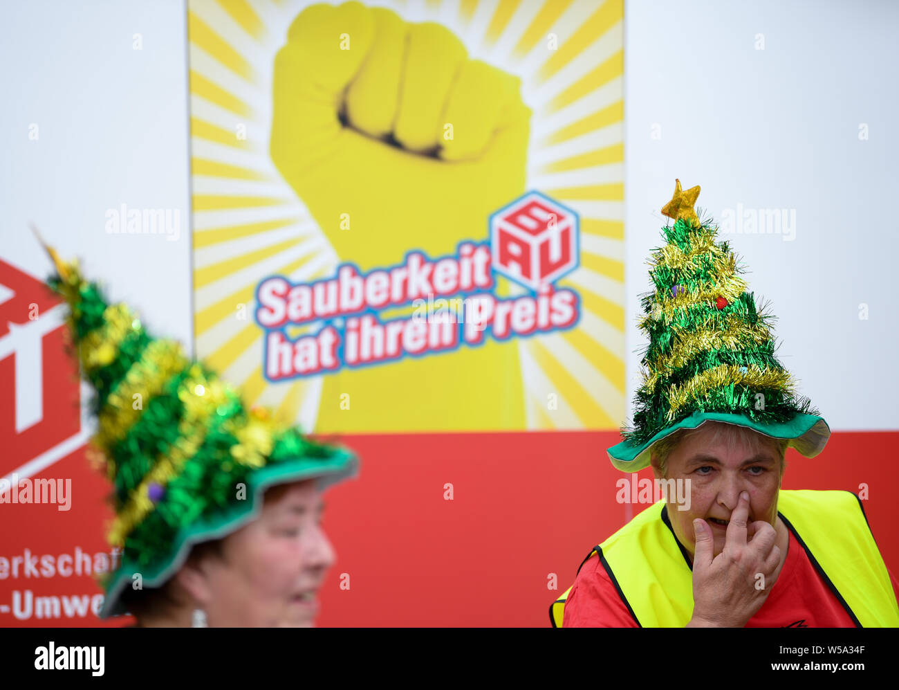 27 July 2019, Hessen, Gießen: With Christmas tree hats on their heads, two women stand in front of a poster with the inscription 'Sauberkeit hat ihren Preis' (Cleanliness has its price) at the action day of the Industriegewerkschaft Bauen-Agrar-Umwelt (IG BAU). Under the motto 'politicians clean', the union is demonstrating against grievances in the cleaning industry. According to the IG BAU, Hesse's 73,000 cleaners face an uncertain future because the employers have terminated the collective agreement for the industry. Photo: Arne Dedert/dpa Stock Photo