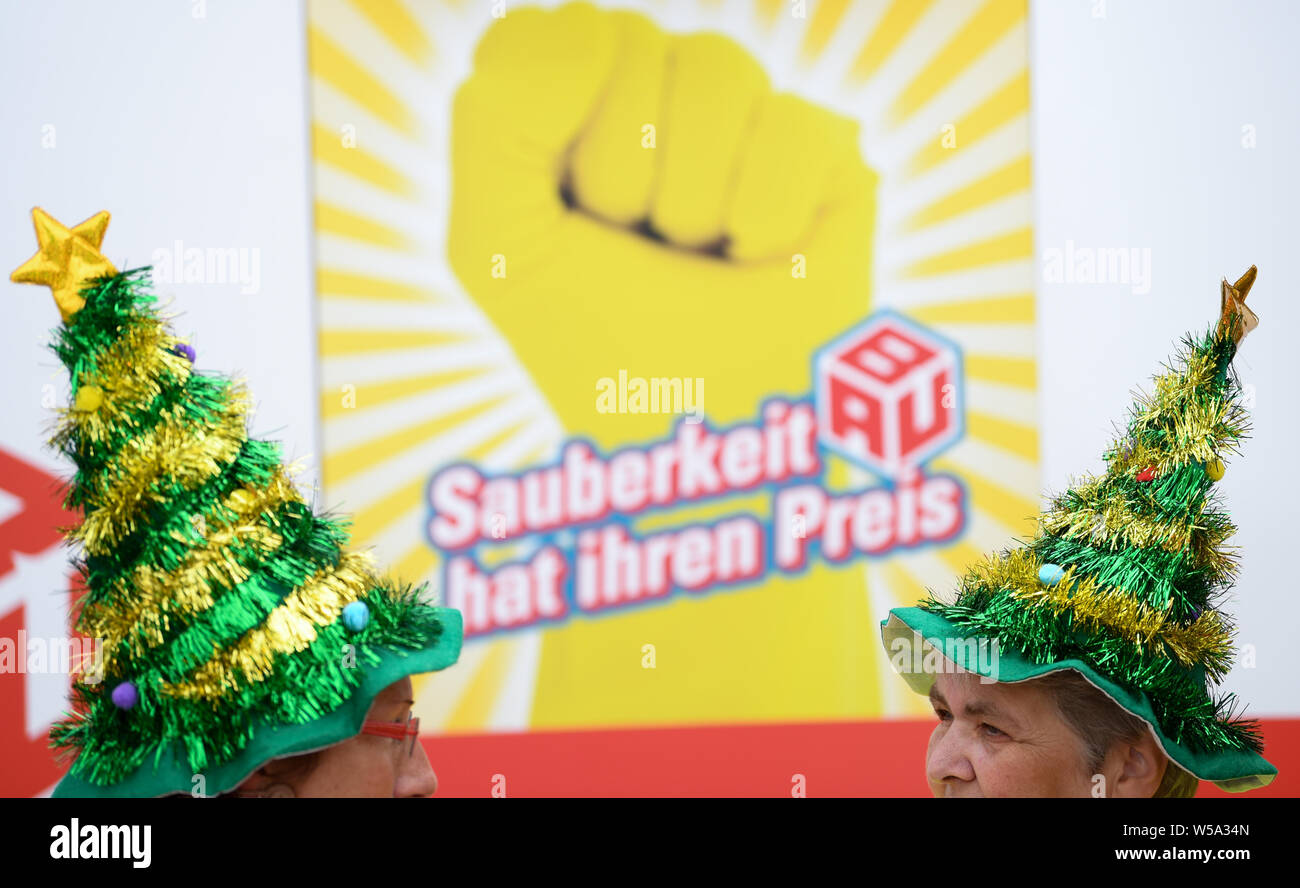 27 July 2019, Hessen, Gießen: With Christmas tree hats on their heads, two women stand in front of a poster with the inscription 'Sauberkeit hat ihren Preis' (Cleanliness has its price) at the action day of the Industriegewerkschaft Bauen-Agrar-Umwelt (IG BAU). Under the motto 'politicians clean', the union is demonstrating against grievances in the cleaning industry. According to the IG BAU, Hesse's 73,000 cleaners face an uncertain future because the employers have terminated the collective agreement for the industry. Photo: Arne Dedert/dpa Stock Photo