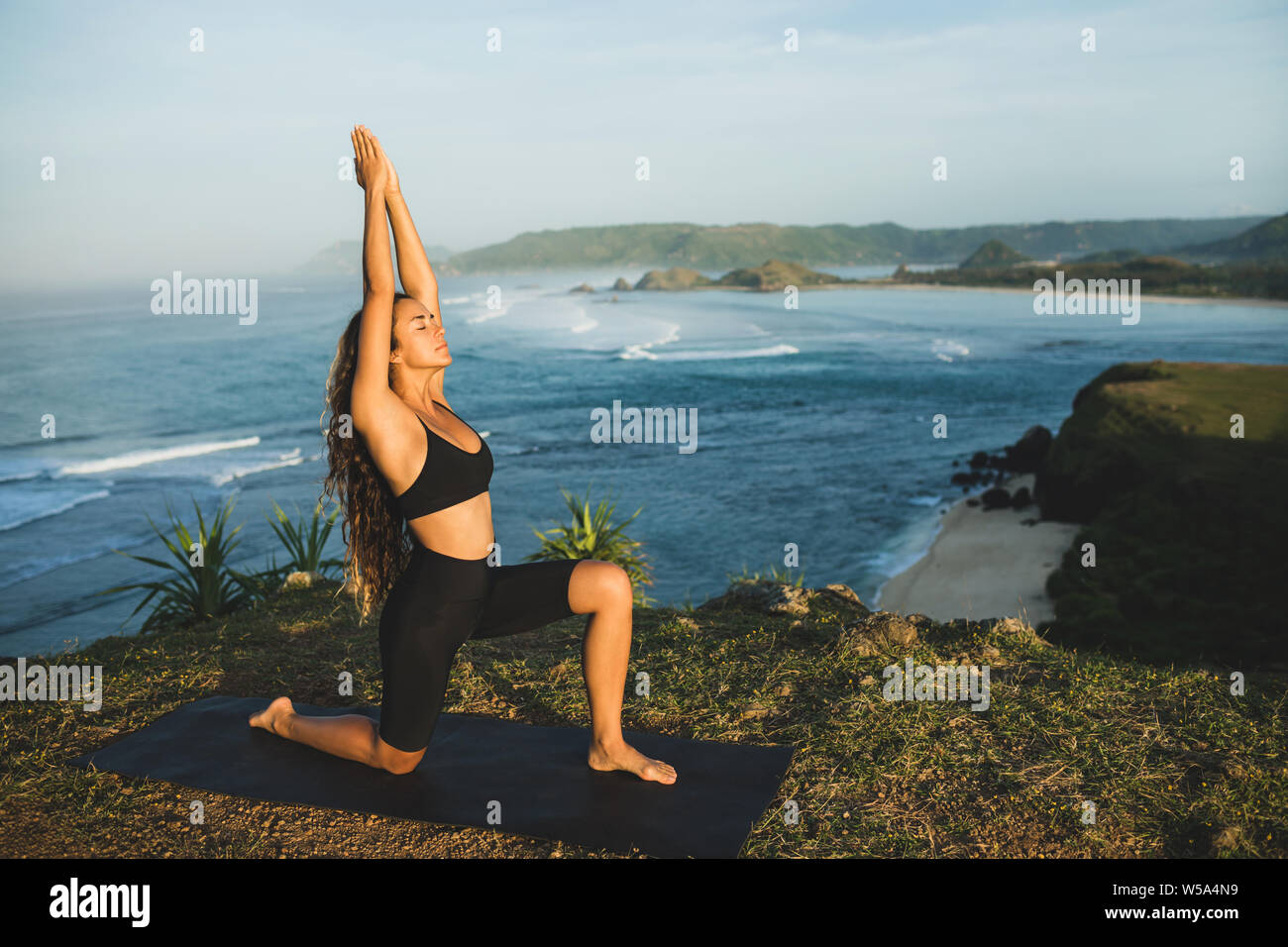 Woman practicing yoga outdoors with amazing ocean and mountain view in morning. Female health and fitness concept. Nature background. Stock Photo