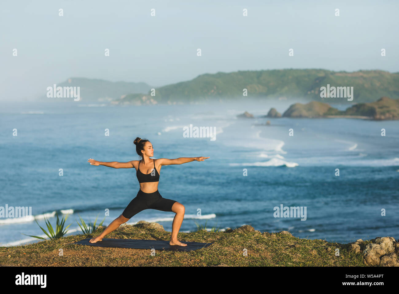 Woman practicing yoga outdoors with amazing ocean and mountain view in morning. Female health and fitness concept. Nature background. Stock Photo