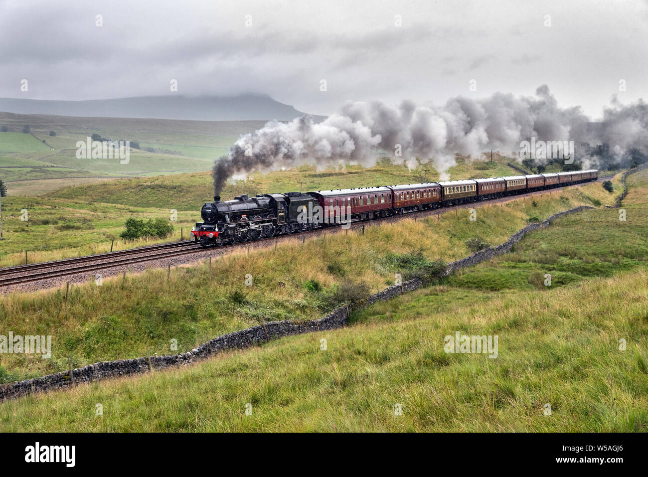 Ribbesdale, North Yorkshire, UK. 27th July, 2019. Climbing the steep gradient on the famous Settle-Carlisle railway line on a wet day in the Yorkshire Dales, steam locomotive 'Leander' with 'The Hadrian' special is seen at Selside, near Horton-in- Ribblesdale. Pen-y-ghent peak is seen under cloud on the horizon. Credit: John Bentley/Alamy Live News Stock Photo