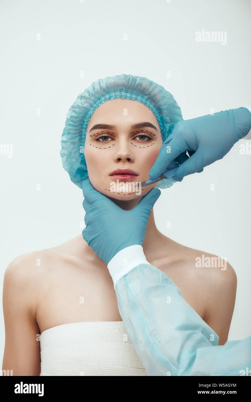 Anti aging treatment. Beautiful young woman in blue medical hat with dashed lines on her face having cosmetic face surgery plastic surgery. Plastic surgeon in blue gloves holding scalpel. Beauty concept. Stock Photo