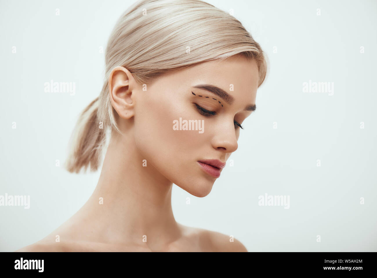 Before facial surgery. Side view of pretty young blonde woman with sketch on her face standing against grey background. Plastic surgery concept. Healthcare. Beauty concept. Stock Photo